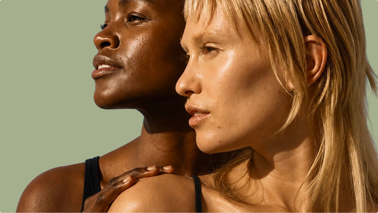 Two women in close profile pose against a muted sage-green background, one resting her hand on the other's shoulder.