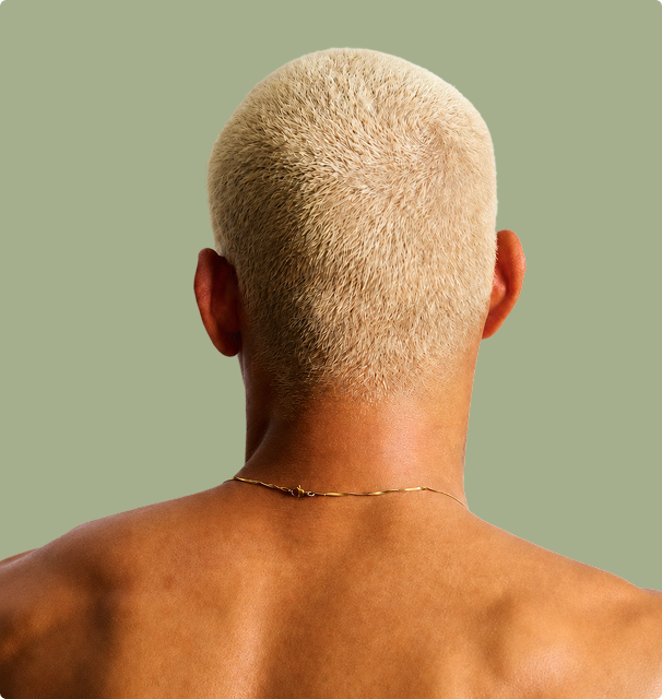 Back view of a person with closely cropped blond hair, a gold necklace, and bare shoulders against a muted green background.